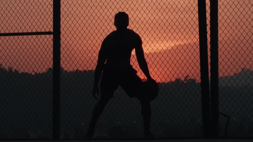 Silhouette of a man dribbling a basketball in slow motion on an outdoor court at sunset