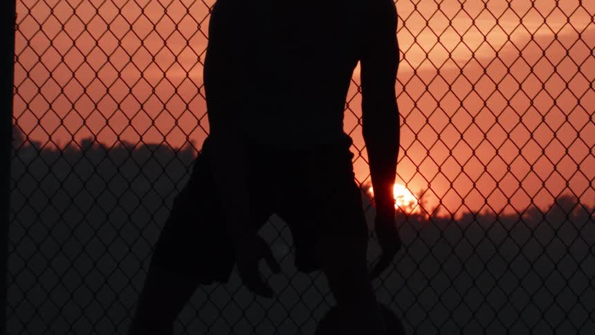 Silhouette of a basketball player dribbling in slow motion in front of a chain-link fence as the sun sets