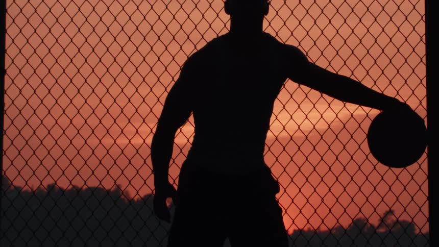 A powerful silhouette of a muscular basketball player holding a ball against a chain-link fence as the sun sets in a dramatic orange sky