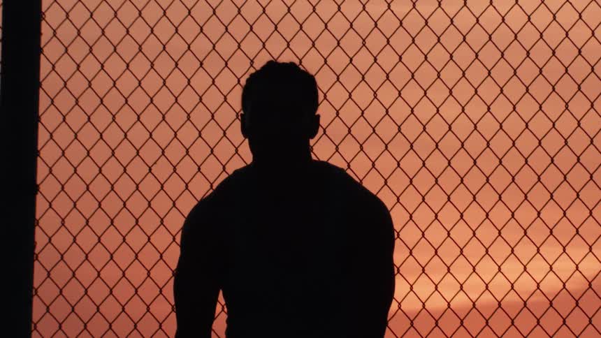 Silhouette of a man with a basketball standing by a chain-link fence at sunset in slow motion