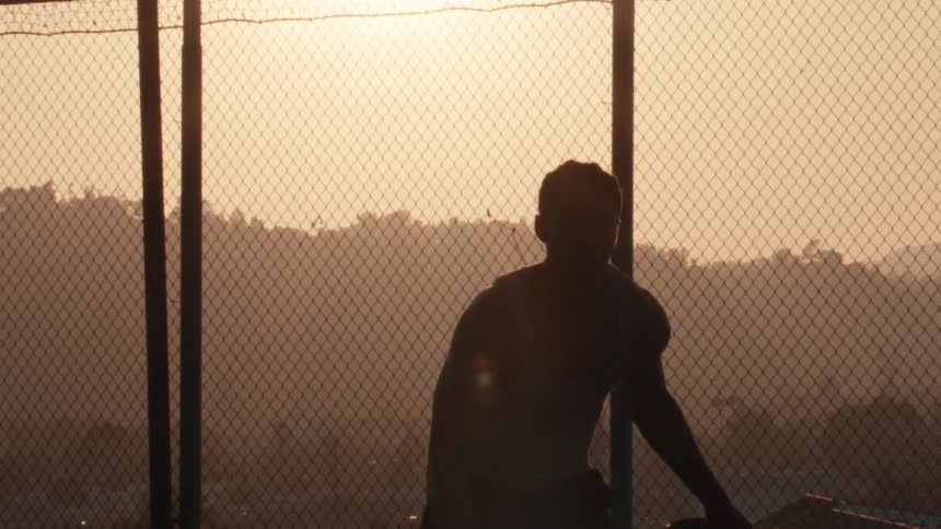 A silhouetted basketball player stands powerfully on an urban court at sunset, seen through a chain-link fence in slow motion