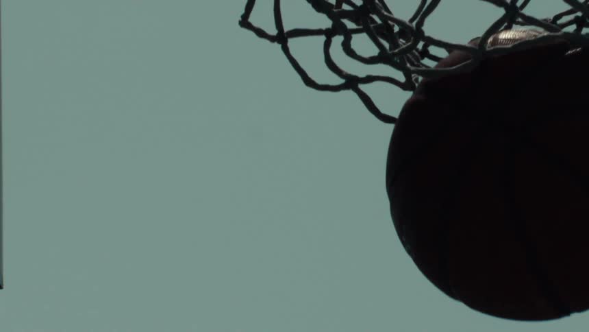 A silhouetted basketball swishes through the net in super slow motion against an overcast sky