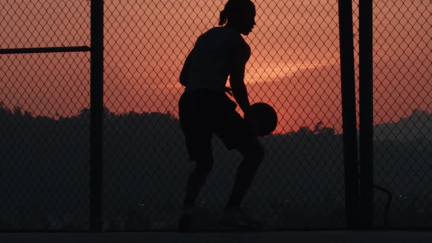 A silhouette of a person dribbling a basketball in slow motion on an outdoor court against a vibrant sunset sky