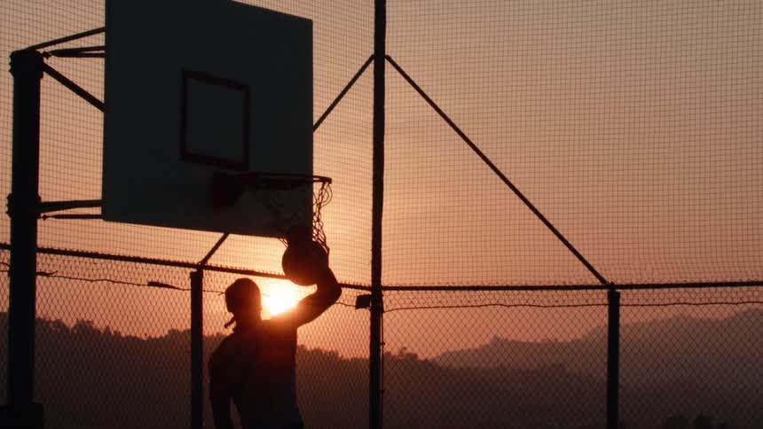 Dramatic slow-motion silhouette of a basketball player jumping and dunking the ball on an outdoor court as the sun sets behind the hills