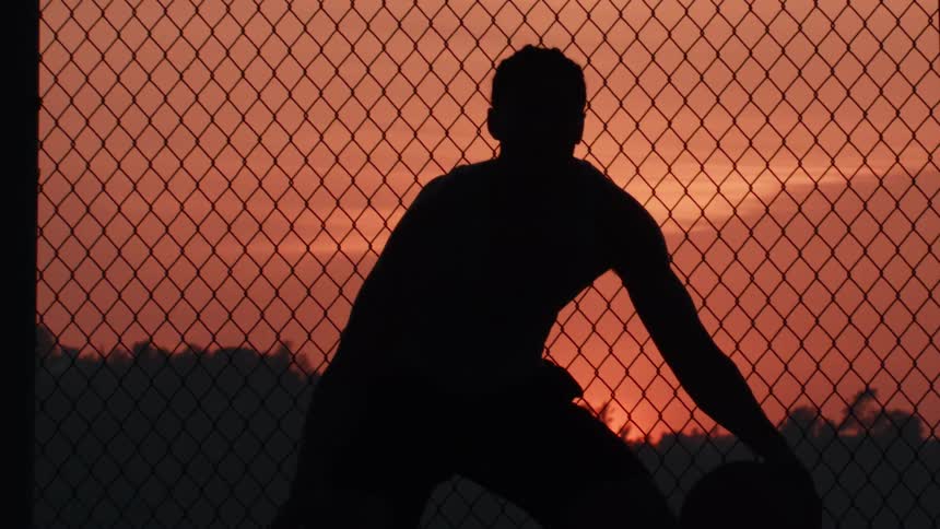 A silhouette of a muscular basketball player dribbling a ball in slow motion in front of a chain-link fence during a beautiful sunset