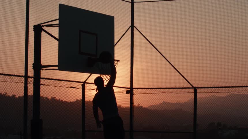 A silhouetted basketball player leaps through the air to perform a powerful slam dunk in slow motion against a beautiful sunset