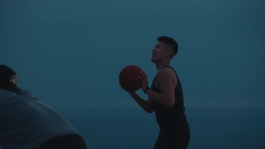 A young man shoots a basketball in slow motion as the twilight sky sets in over the ocean