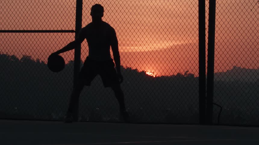 Silhouette of a man dribbling a basketball on an outdoor court as the sun sets in the background