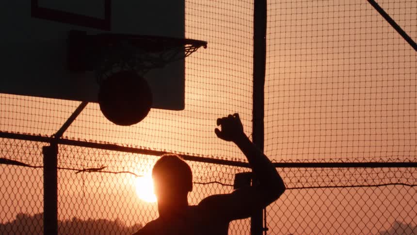 Slow motion silhouette of a basketball player executing a powerful slam dunk against the golden sunset