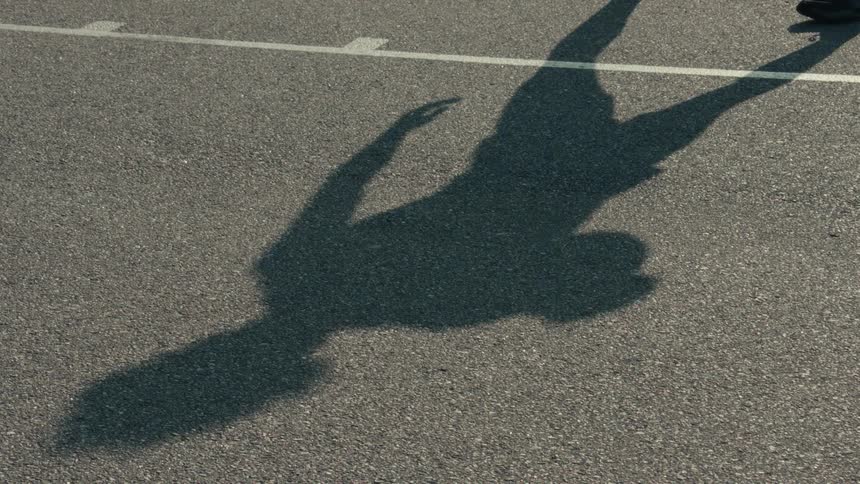 The long shadow of a basketball player is cast on the asphalt court as they dribble the ball in slow motion