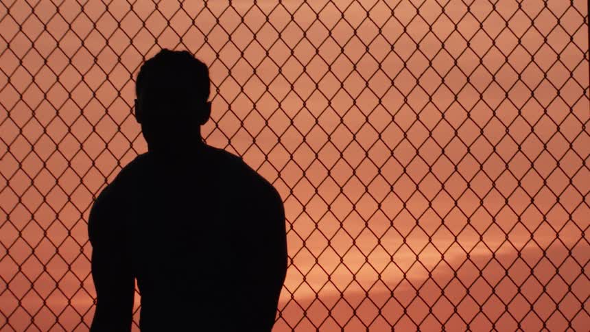 Silhouette of a lone basketball player standing by a chain-link fence court during a beautiful orange sunset