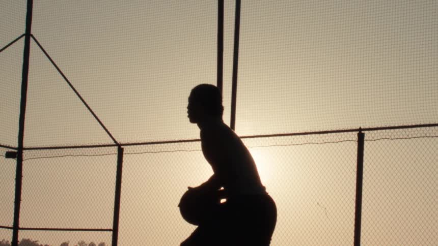 A basketball player's silhouette is captured in slow motion as they practice their free throw shot on an outdoor court during a golden sunset