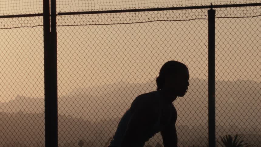 A female basketball player stands silhouetted against the golden sunset on an urban court with mountains in the background
