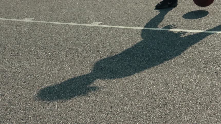 A basketball player's shadow is cast on the asphalt court as they dribble a ball in super slow motion