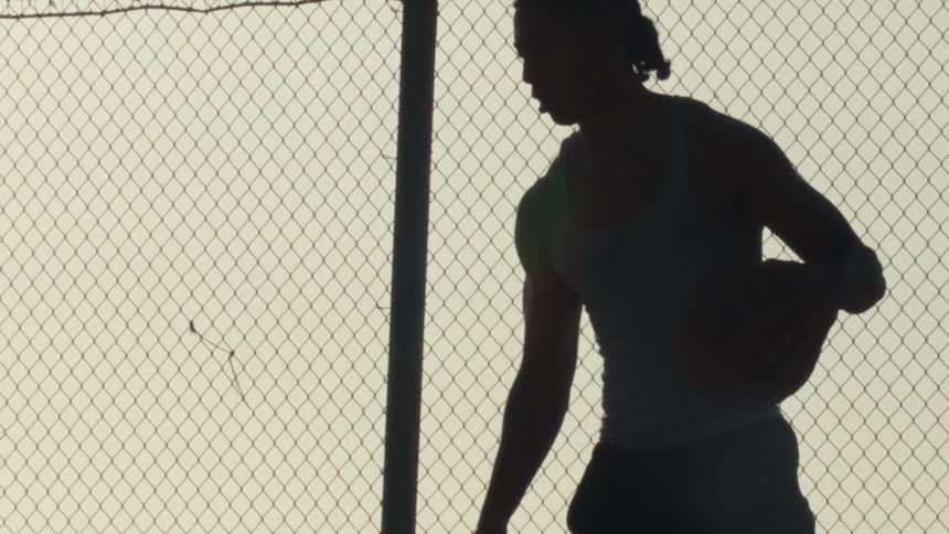 A silhouette of an athletic man dribbling a basketball in slow motion against a chain-link fence at sunrise