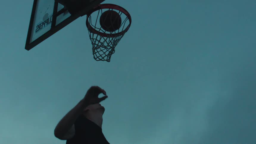 Low angle slow-motion shot of a man's silhouette as he jumps up and slam dunks a basketball at twilight