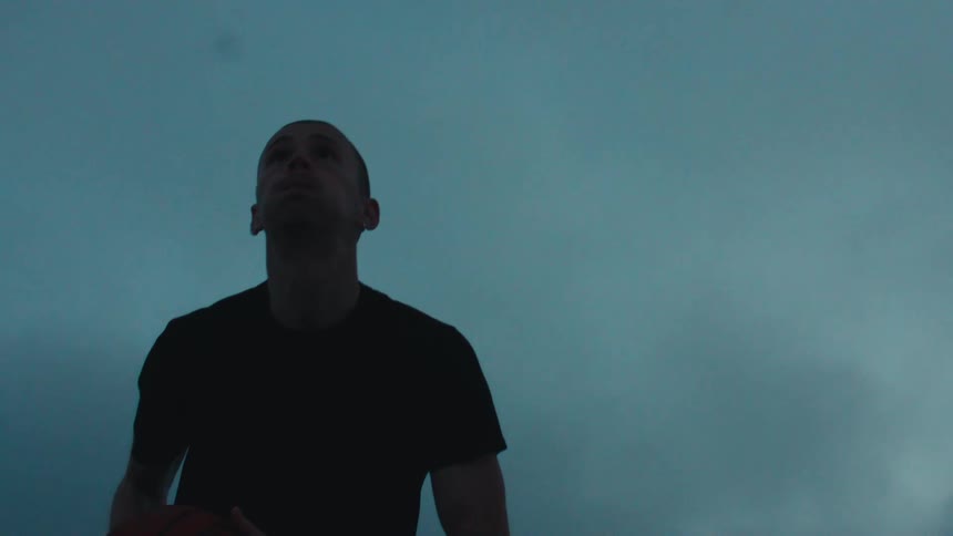 A focused male basketball player dribbles and then shoots a basket against a moody, dark sky in dramatic slow motion