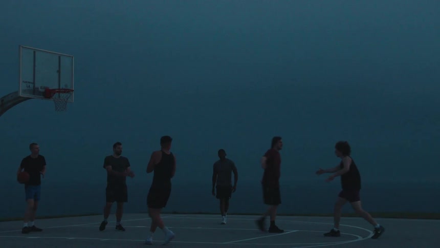 A young man makes a perfect jump shot in slow motion while playing basketball with his friends on a moody, atmospheric court at dusk