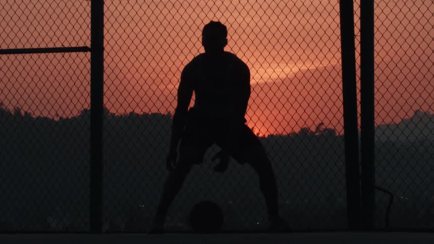 Slow-motion silhouette of a basketball player dribbling a ball on a fenced-in court against a vibrant sunset sky