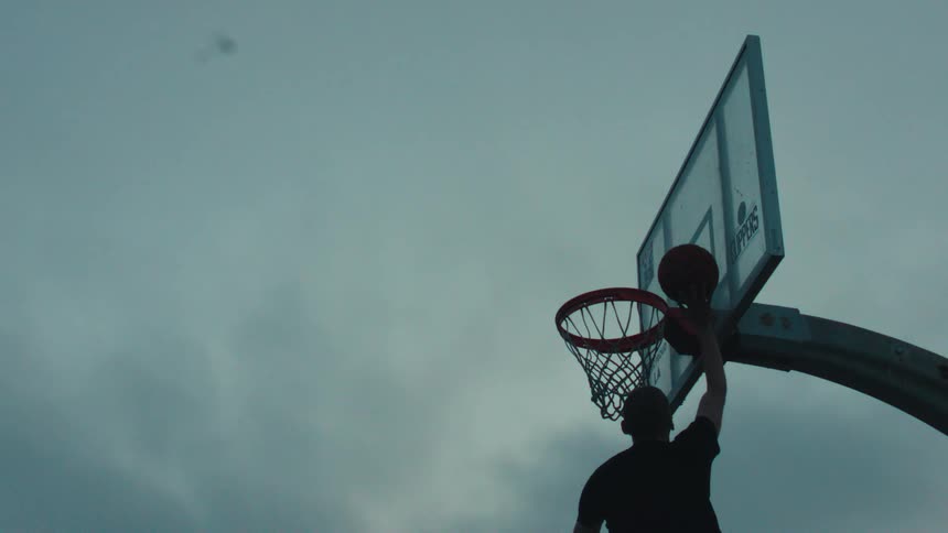 A silhouetted basketball player leaps toward the hoop and scores a basket in super slow motion against a dramatic overcast sky