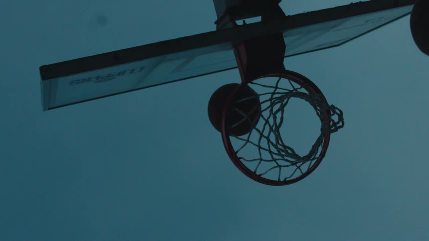 From a low angle, a silhouetted basketball player scores a basket in dramatic slow motion against the twilight sky