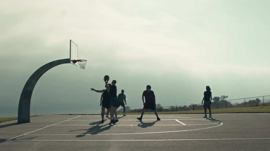 Friends play a game of pickup basketball on an outdoor court in slow motion, with one player sinking a contested jump shot