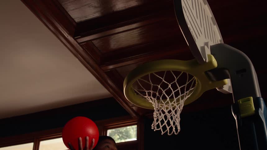 A young boy practices his slam dunk in slow motion on a toy basketball hoop in a dimly lit room