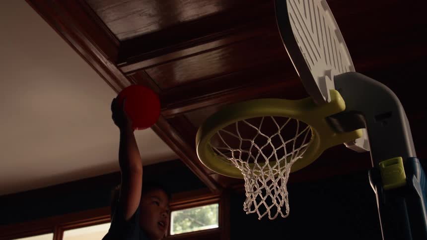 A young Asian boy slam dunks a red ball into a toy basketball hoop inside his house in slow motion