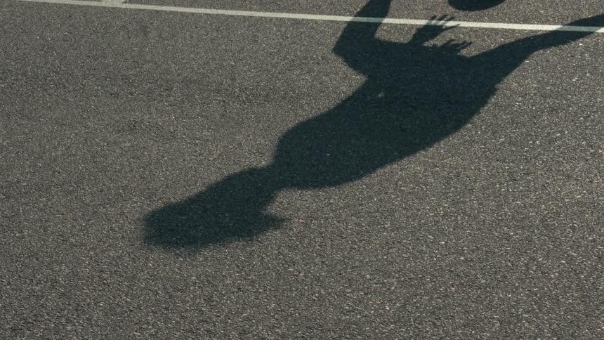 The silhouette of a basketball player is cast on the pavement as they practice dribbling in slow motion