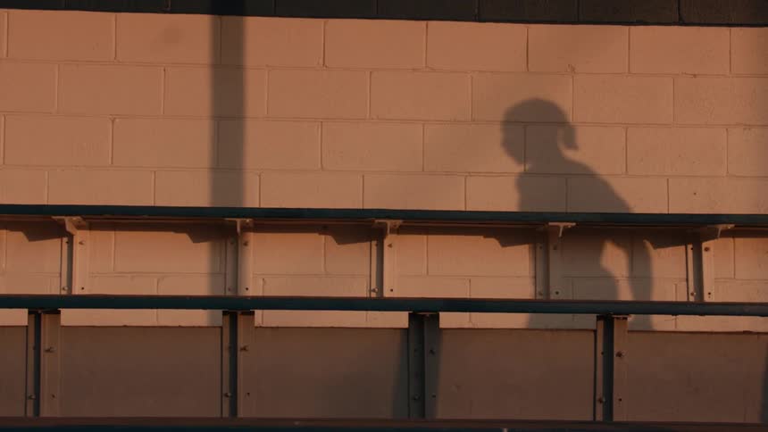 The shadow of a basketball player is cast on a white brick wall behind empty bleachers during golden hour