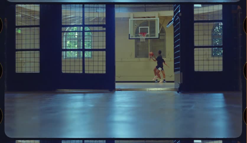 A young man and woman play one-on-one basketball on an indoor court as the man makes a successful shot in slow motion