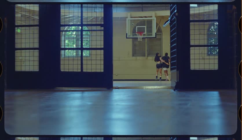 A man scores a basket while playing a one-on-one game with a woman on a moody indoor court, framed through a large doorway