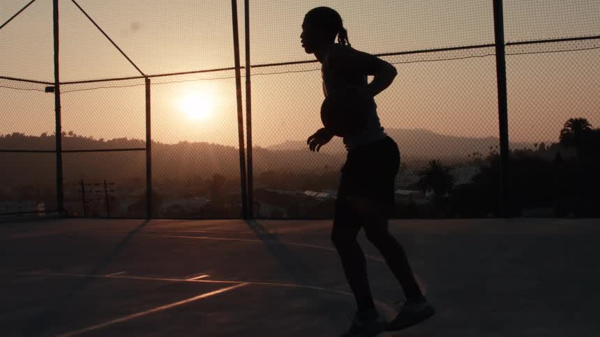 A silhouetted basketball player dribbles a ball in slow motion on an outdoor court at sunset