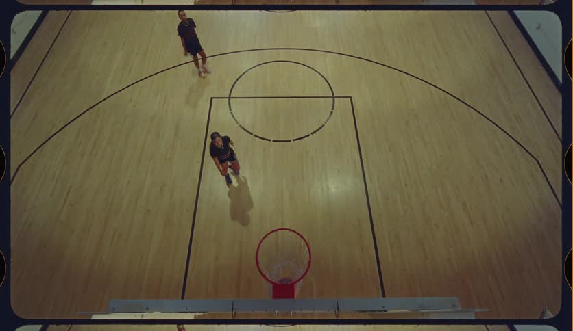 A high-angle, birds-eye view shows a man and a woman playing one-on-one basketball on an indoor court, with the man making a successful shot