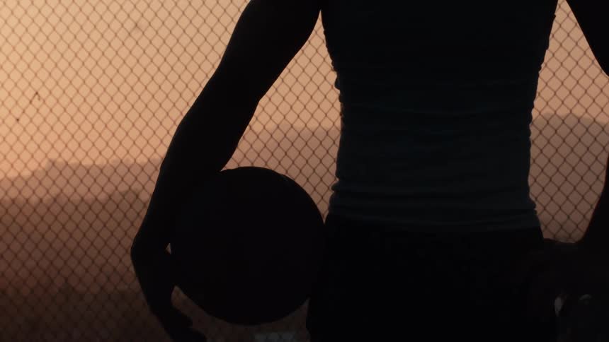 A silhouetted basketball player stands thoughtfully holding a ball against a chain-link fence during a golden sunset