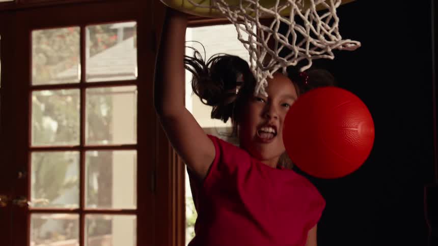A determined young girl with pigtails dunks a red ball into an indoor basketball hoop in super slow motion