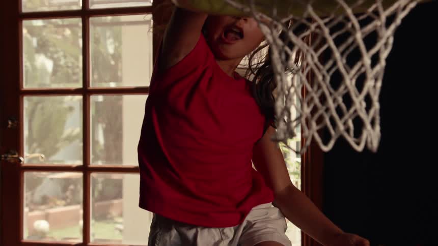 Focused young girl in a red shirt makes an impressive slam dunk on her indoor basketball hoop in dramatic slow motion