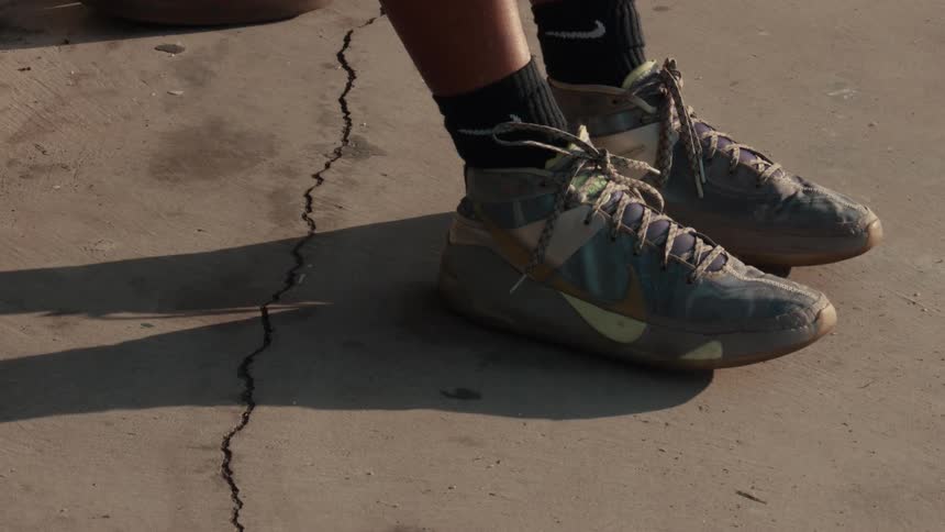 A person in basketball sneakers sits on a court and slowly dribbles a basketball in a close-up, slow-motion shot