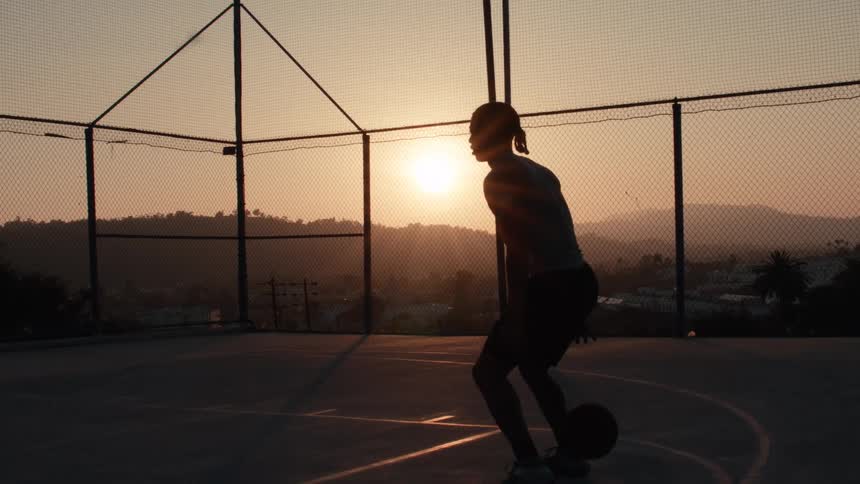 Cinematic slow motion shot of a basketball player dribbling on a court silhouetted by the golden hour sunset