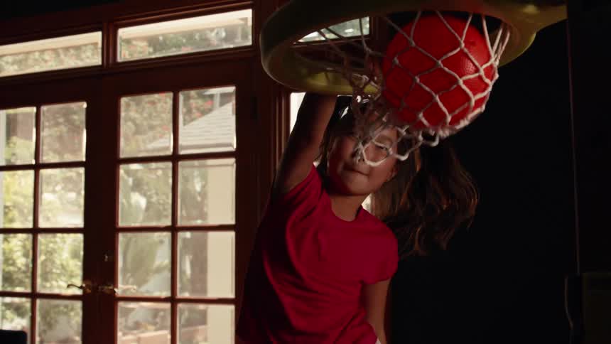 A young girl in a red shirt with pigtails happily slam dunks a toy basketball into an indoor hoop in slow motion