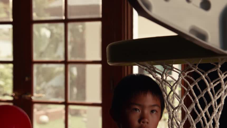A young boy rises up in slow motion to slam dunk a red toy basketball and winks triumphantly