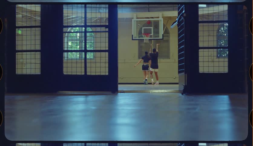 Viewed through a large open doorway, two friends play basketball on an indoor court, and one scores a basket