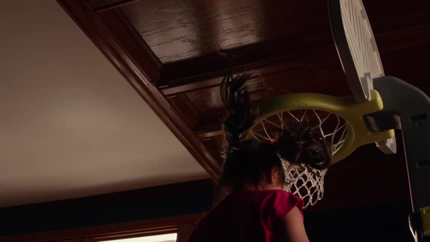 A young girl slam dunks a red toy basketball into an indoor hoop in slow motion