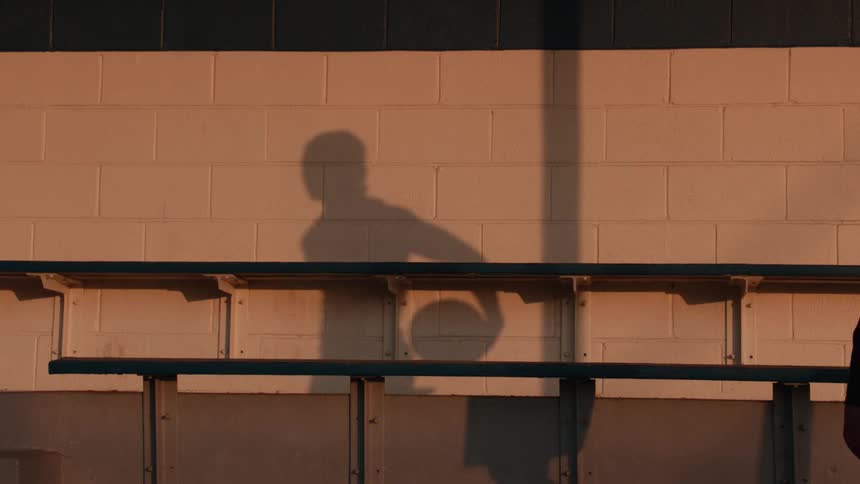 The shadow of a man holding a basketball is cast on a brick wall as he walks past in slow motion at golden hour