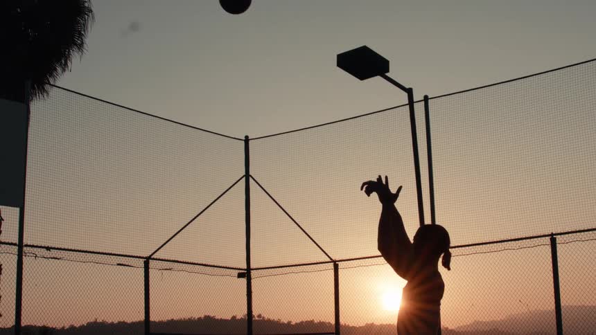 Slow motion silhouette of a person shooting a basketball on an outdoor court during a golden sunset