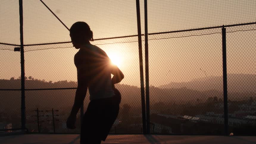 Slow motion silhouette of an athletic man practicing basketball on an outdoor court at sunset