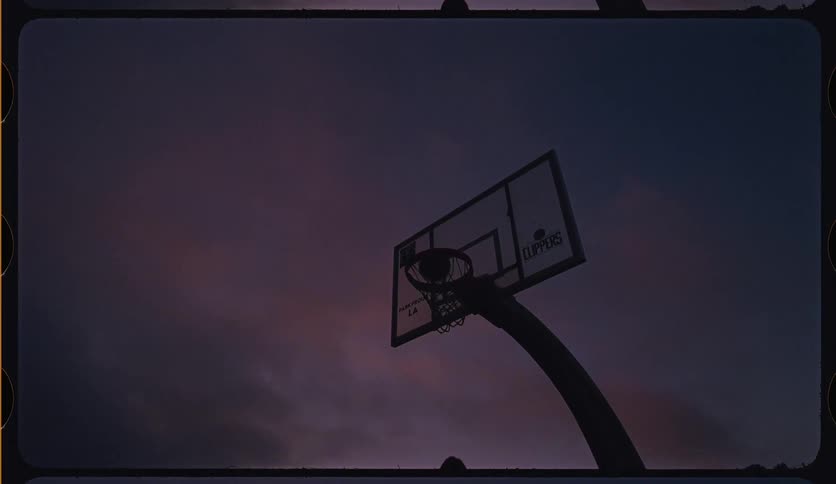 A silhouetted basketball player shoots and scores a layup in slow motion against a dramatic twilight sky
