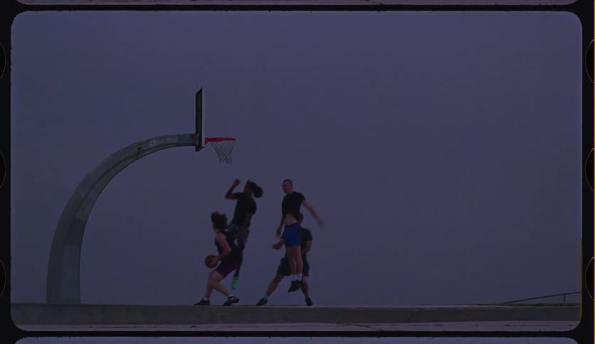 Friends play a dramatic slow-motion basketball game on an outdoor court at dusk