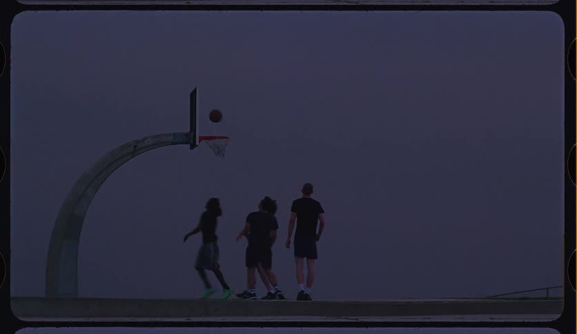 Four young men are silhouetted against a dark sky as they play a game of pickup basketball in super slow motion on an outdoor court
