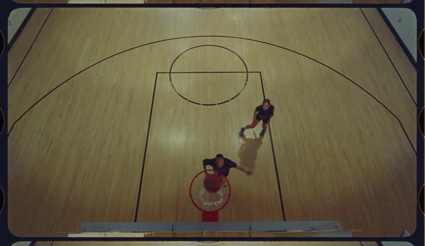 From a high angle, a woman skillfully dribbles past a defender to score a layup in slow motion on an indoor basketball court
